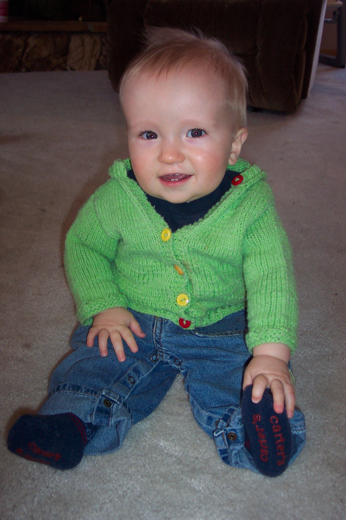 A baby boy sitting up and smiling while wearing a lime green hooded knit cardigan.