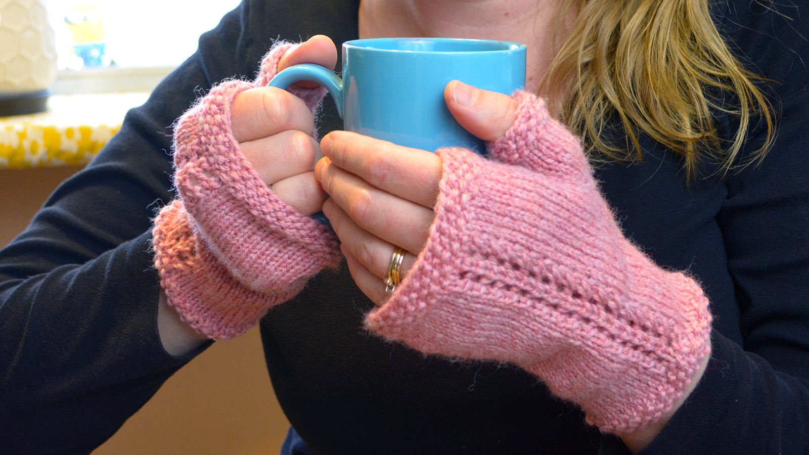A woman’s hands wearing knit fingerless mittens and holding a mug.