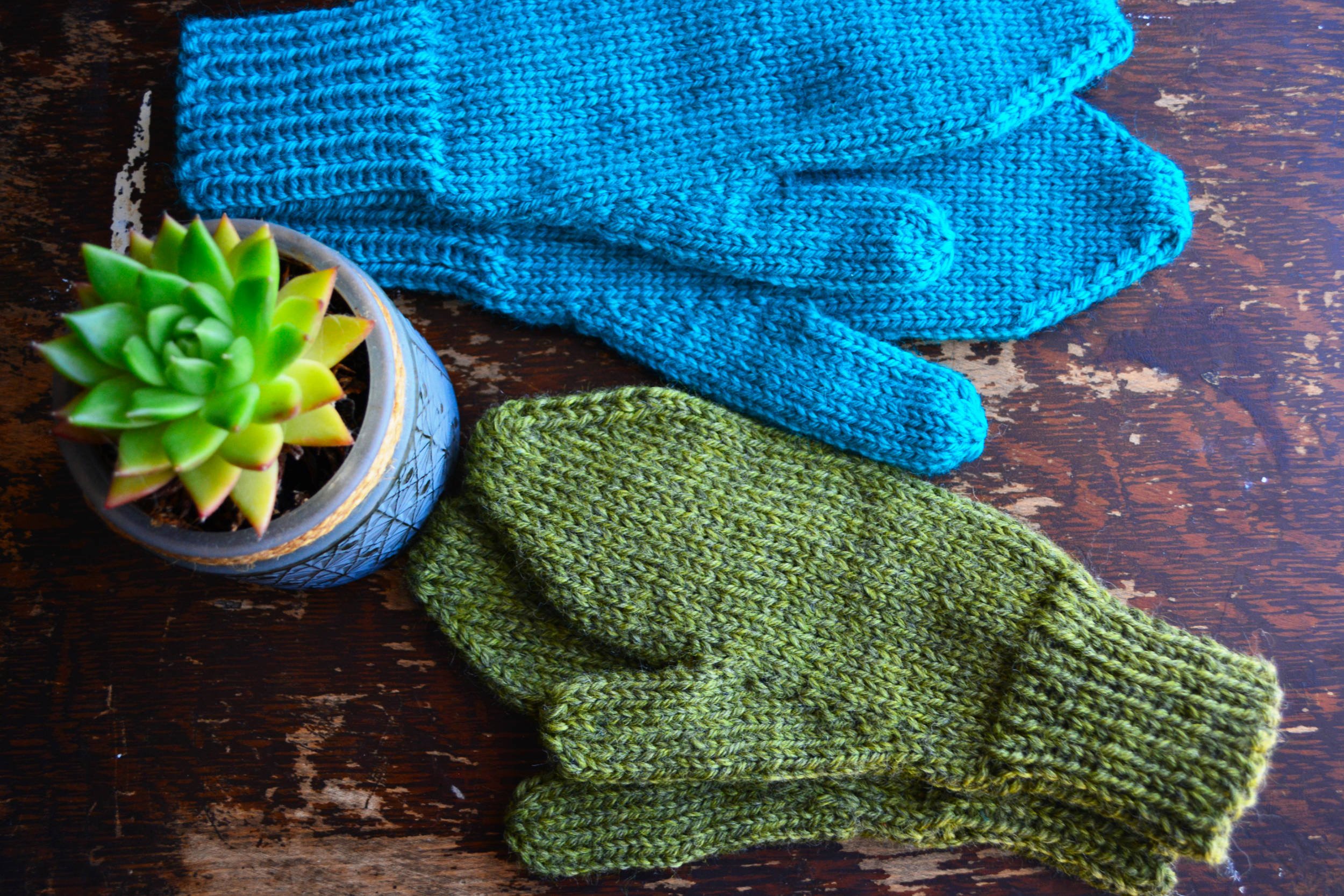 Two pairs of mittens lay on a worn wooden surface next to a spiky succulent. The mittens have gussets, angled tips to a blunt end, and long ribbed cuffs. The thumbs are straight.