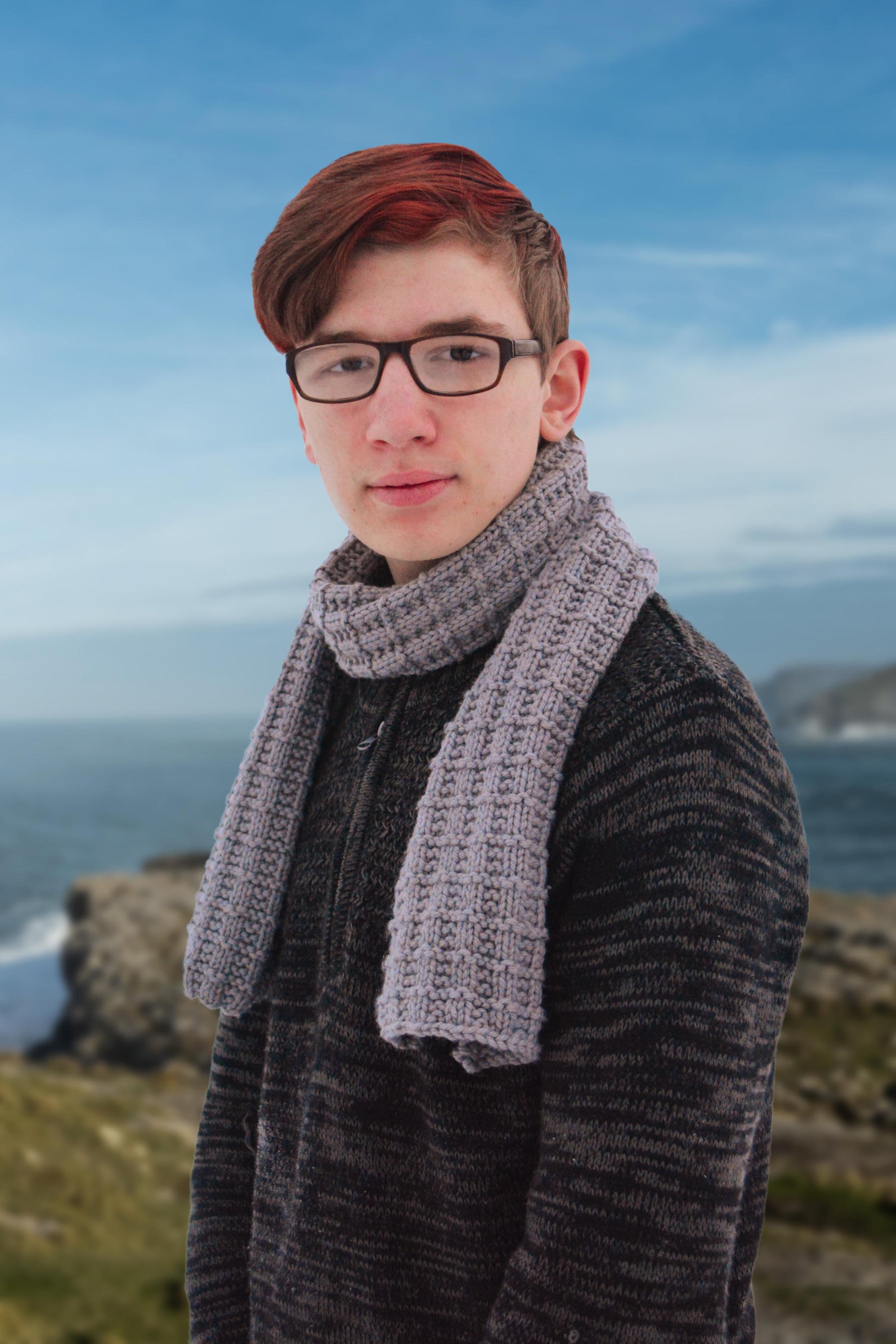 A young man wears a masculine-looking bulky-knit scarf with waffle weave texture and stands in front of a turbulent sea on a rocky shore.