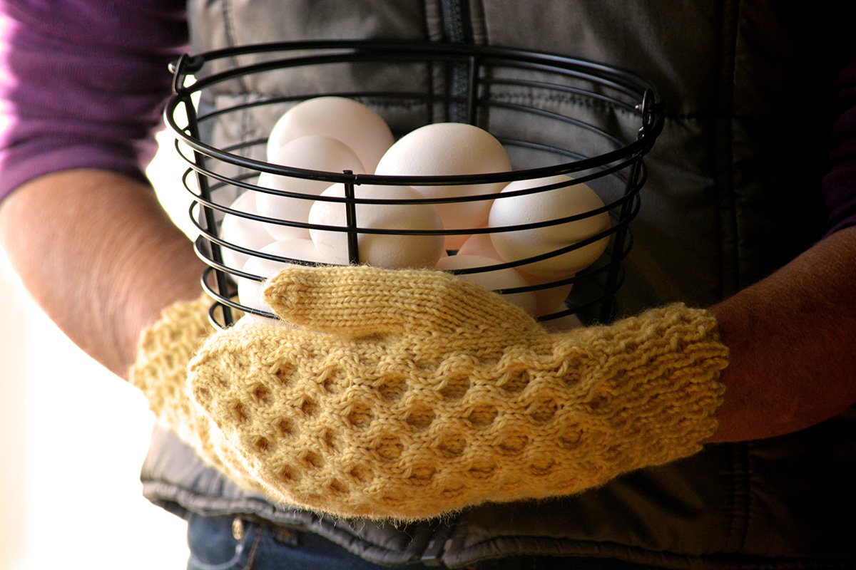 The Honey Tree mittens on a woman's hands holding a basket of eggs.