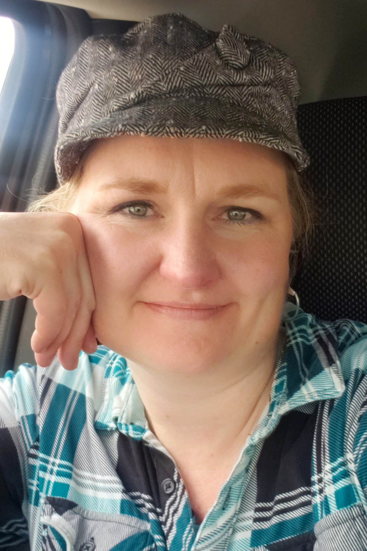 Talena Winters, a blond Caucasian woman, leans her cheek on her hand in a car and smiles at the camera while wearing a fabric newsboy cap and a teal plaid shirt